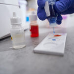 Laboratory assistant in blue gloves standing near table with medical equipment and holding small bottle with blue liquid
